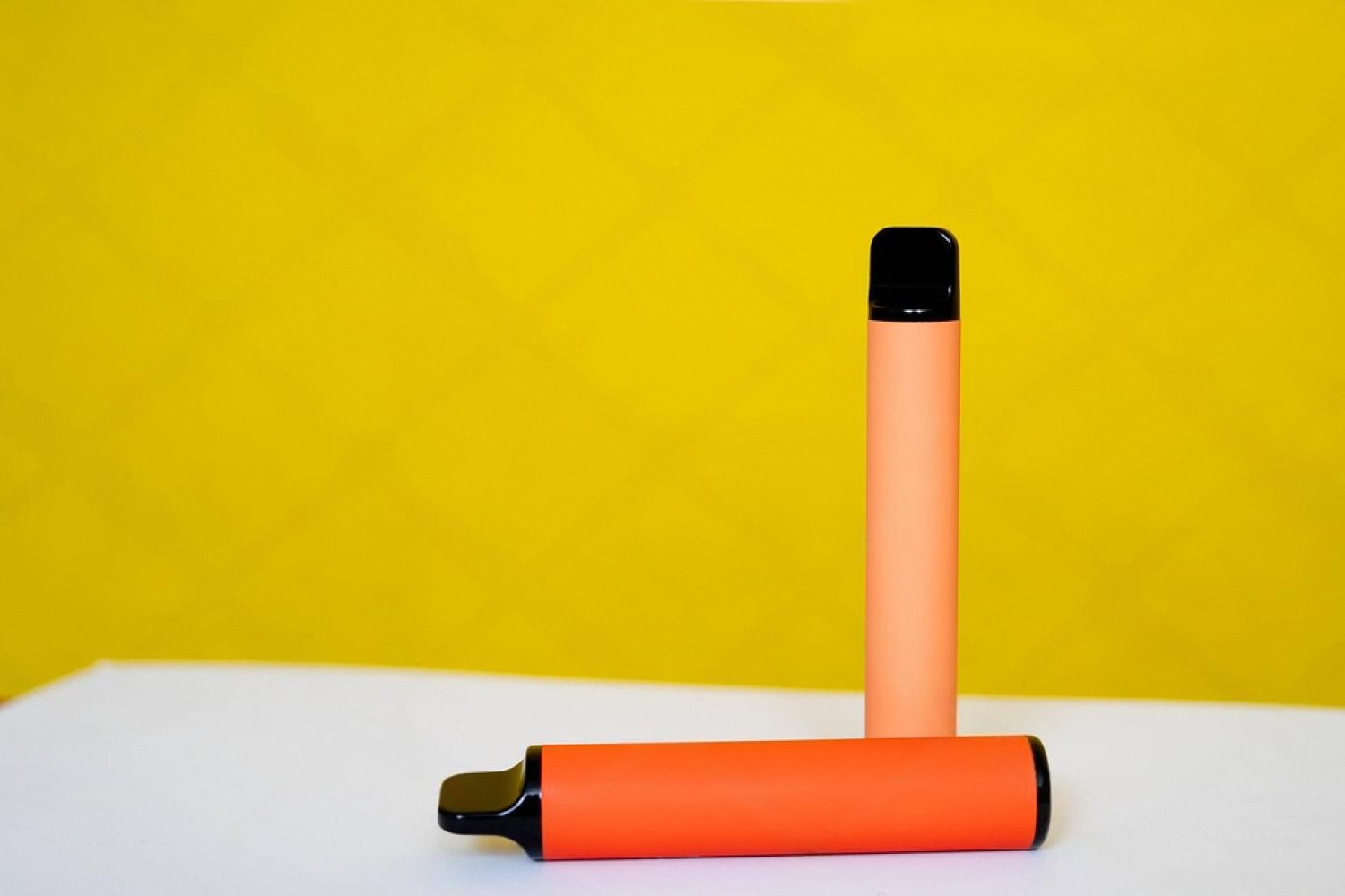 Two orange disposable vape devices on a white surface against a yellow background